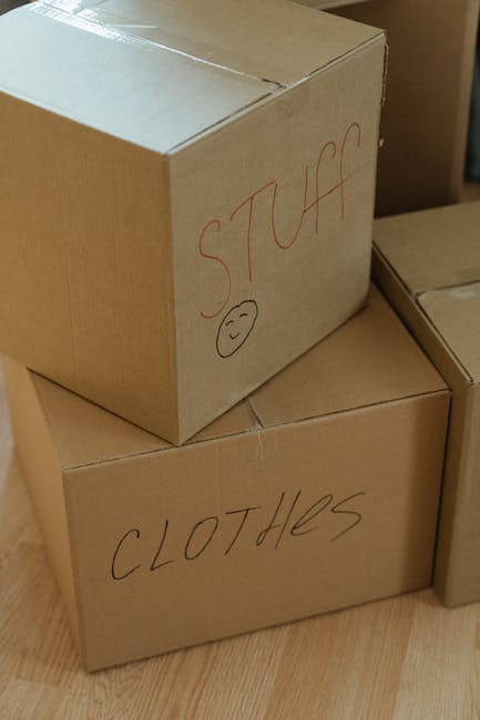 A person dressed in blue work clothes and orange sneakers sitting on a wooden floor inside a room with large arched windows, surrounded by several closed cardboard boxes with red packing tape. The ind