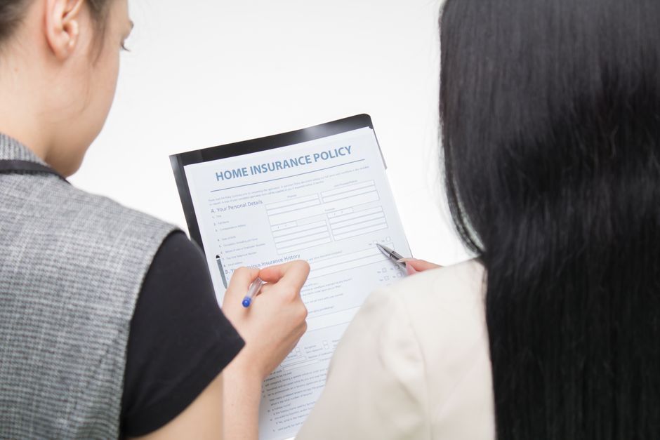 Two women are seated with their backs to the camera, reviewing a document titled 'Home Insurance Policy' printed on white paper with blue text. One woman is wearing a dark grey top with light grey sle