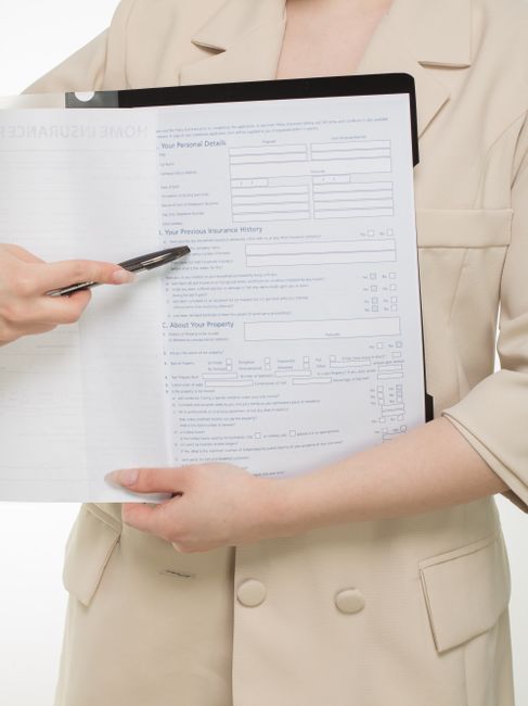 Two women are seated with their backs to the camera, reviewing a document titled 'Home Insurance Policy' printed on white paper with blue text. One woman is wearing a dark grey top with light grey sle