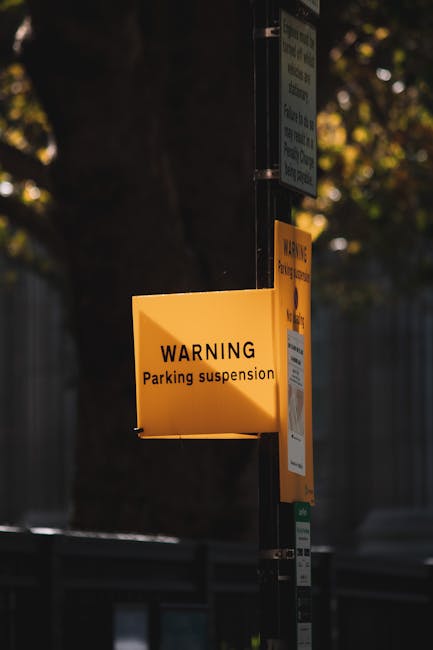 A close-up view of a yellow sign attached to a metal pole, displaying the message 'WARNING Parking suspension' in black text. The sign is positioned outdoors on a street or driveway, with a background